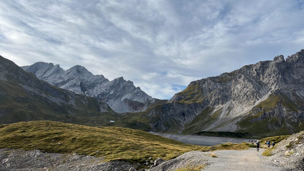 Transalpine Run 2025: Berglandschaft mit Wanderweg und Wanderern unter Wolkenhimmel. - We Run 4 Fun!