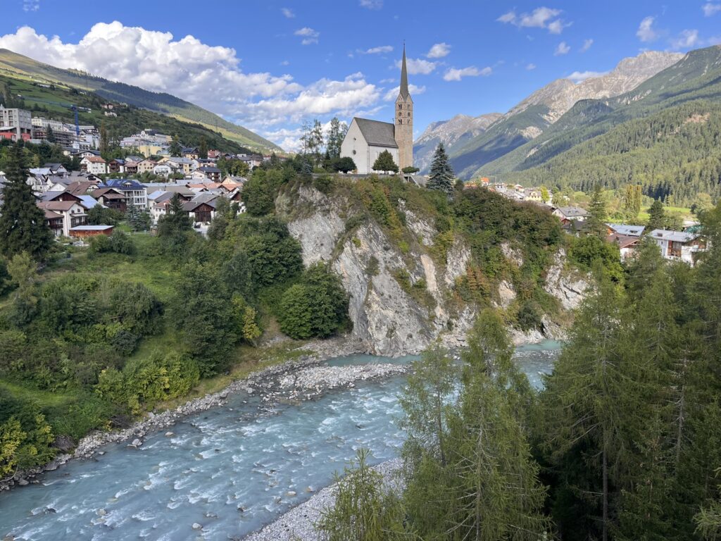 Idyllisches Bergdorf mit Kirche am Flussufer, möglicherweise Teil des Transalpine Run 2025. - We Run 4 Fun!