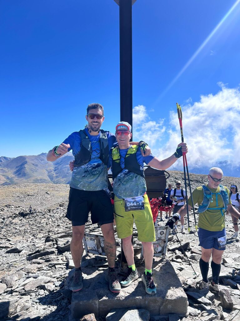 Zwei Läufer auf einem Berggipfel mit Wanderstöcken und Startnummern. - We Run 4 Fun!